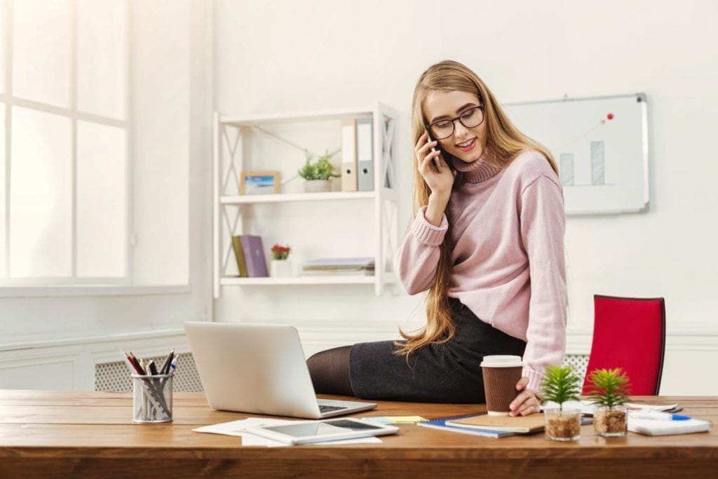 Smiling business woman talking by phone and having coffee, sitting on table at office workplace, copy space. Communication, technology, success concept